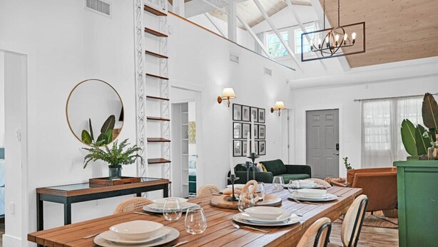 Un coin repas avec une table en bois dressée pour quatre personnes, entourée de chaises en osier. On aperçoit une échelle blanche et une armoire verte à l’arrière-plan.