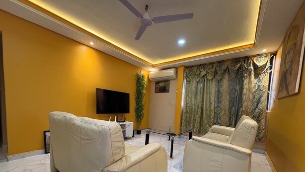 A living room with two beige sofas, a flat-screen TV, a ceiling fan, and a large window with curtains. - Charming Pent house 1-bedroom apartment with kitchen, hall and office for a fee (Accra)