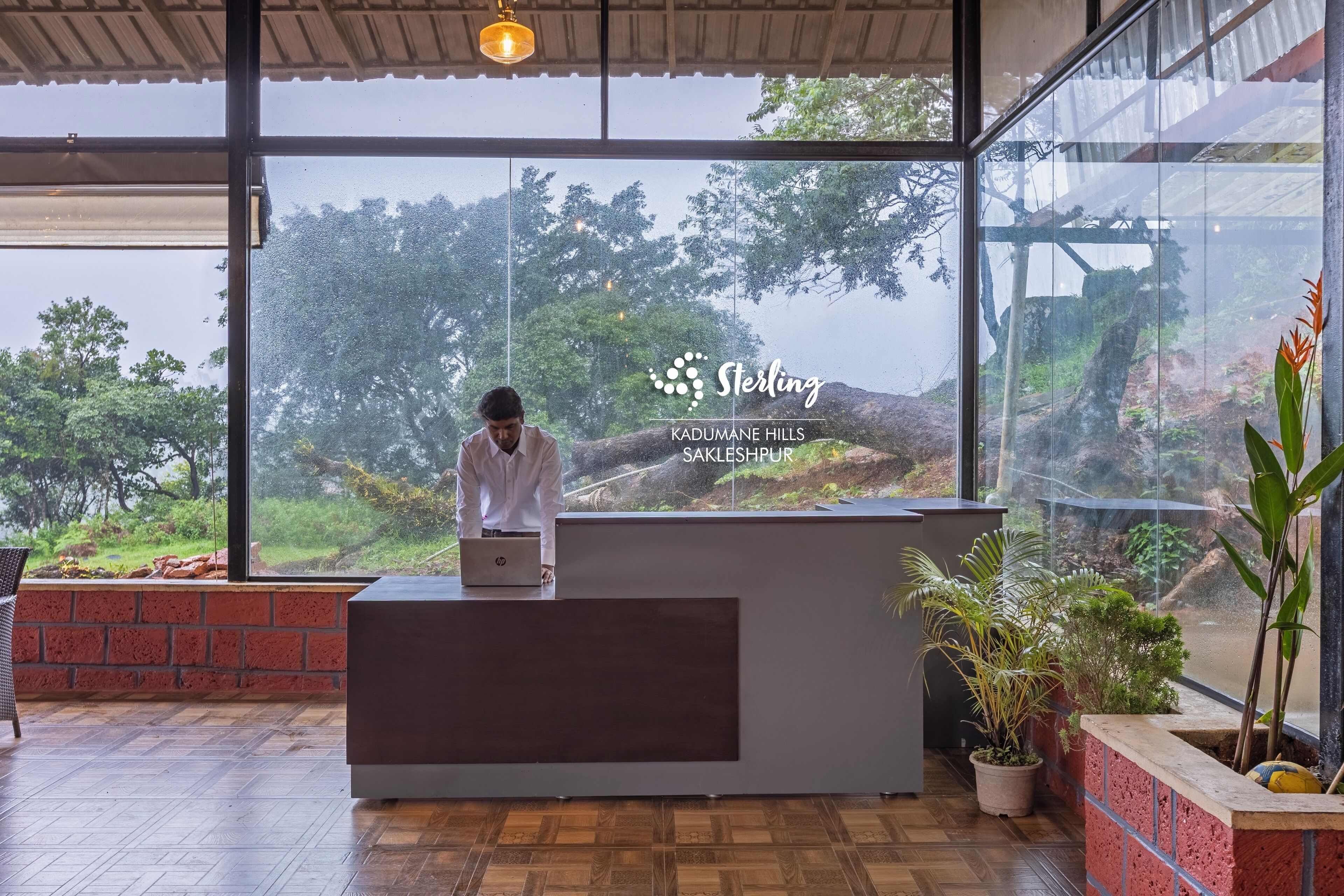A reception desk with a laptop, a large window with a view of Kadumane Hills, and a sign reading 'Sterling Sakleshpur.'