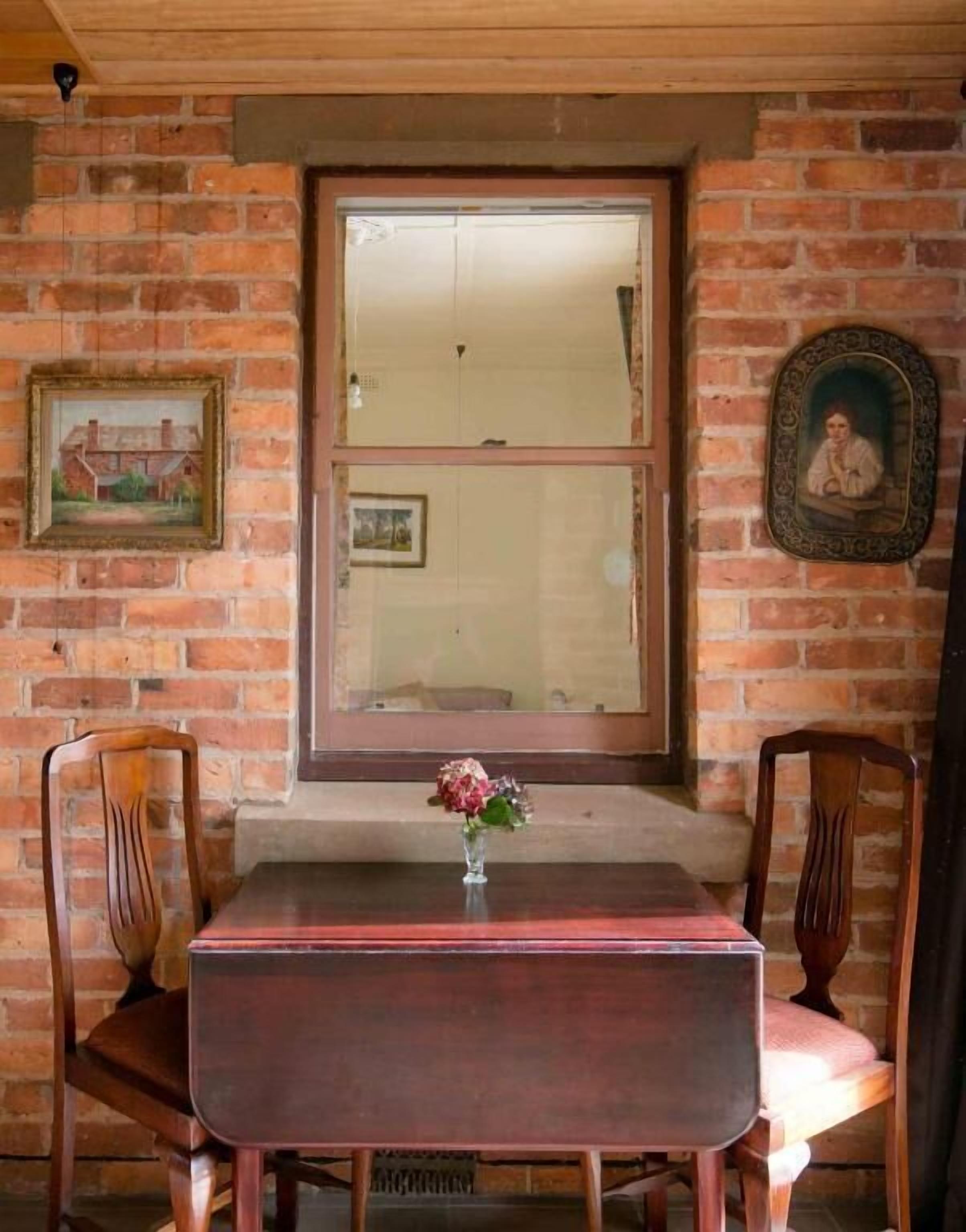 A dining area with a brick wall, wooden chairs, a table, and a vase with flowers.
