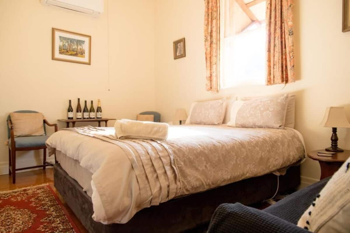 Un dormitorio ordenado con una cama, una silla, una mesita con botellas y una lámpara.