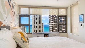 Ein Schlafzimmer mit einem großen Bett, einem Fenster mit Blick auf hohe Gebäude und das Meer sowie einem Schreibtisch mit einer Lampe.