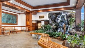Eine Lobby mit Steinfußboden, Holzbänken, einem Wasserfallbrunnen und einem Schild mit der Aufschrift 'Tower 1 Makai'.