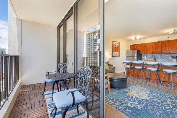 A balcony with a wooden floor, two chairs, a small table, and a glass door leading to a kitchen area with wooden cabinets and a bar counter.
