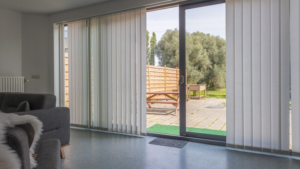 A modern living room with a grey sofa, a furry throw, and a sliding glass door leading to an outdoor patio with a wooden table and chairs. - Charming 2-bedroom villa in brilliant Merelbeke-Melle with AC (Merelbeke-Melle)