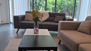A modern living room with a sofa, a coffee table, and a view of greenery through large windows. - Charming 2-bedroom villa in brilliant Merelbeke-Melle with AC (Merelbeke-Melle)
