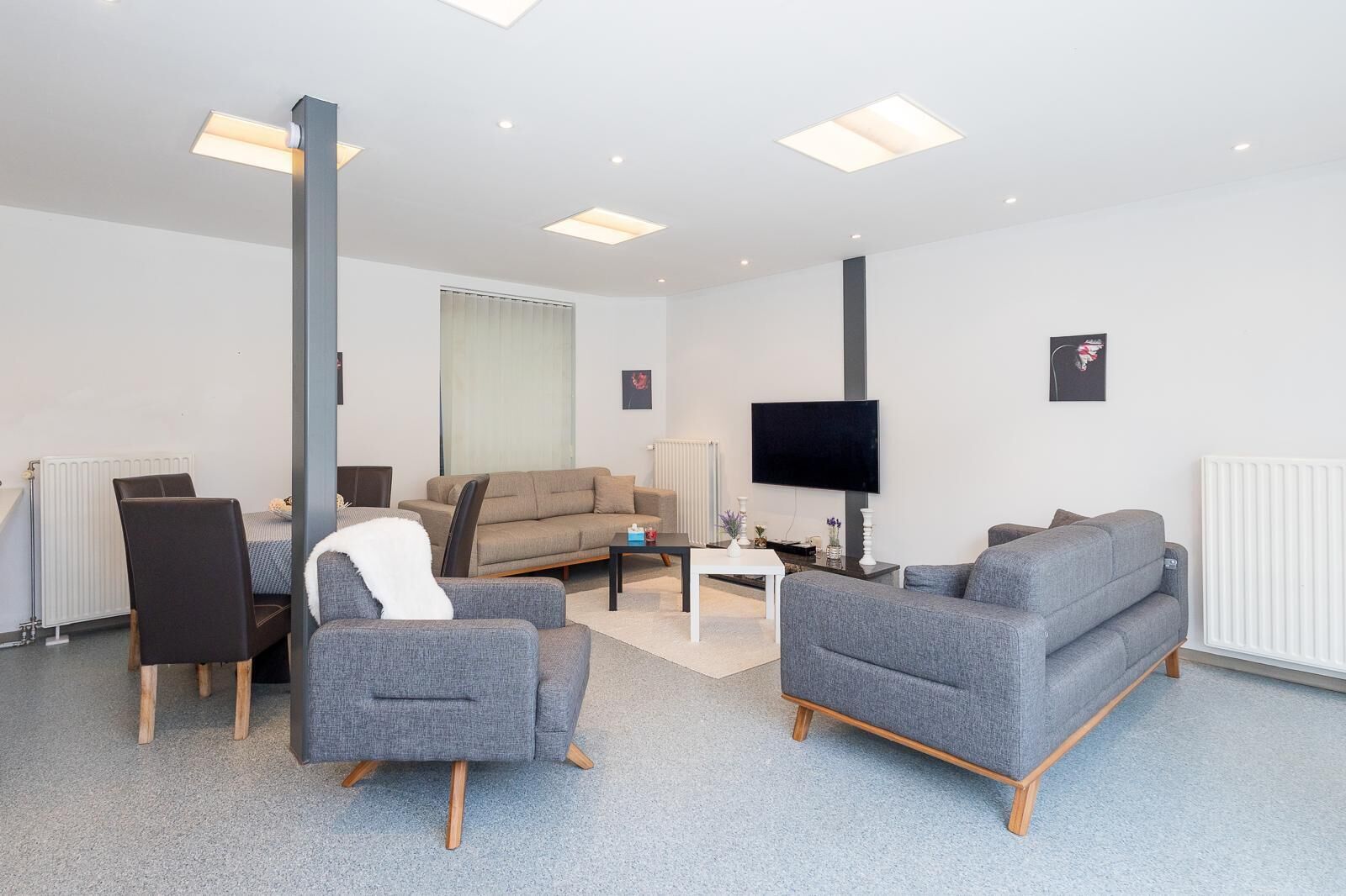 Een moderne woonkamer met een bank, stoelen, een salontafel, een flatscreen-tv en inbouwspots.