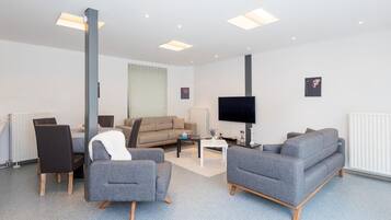 Een moderne woonkamer met een bank, stoelen, een salontafel, een flatscreen-tv en inbouwspots.