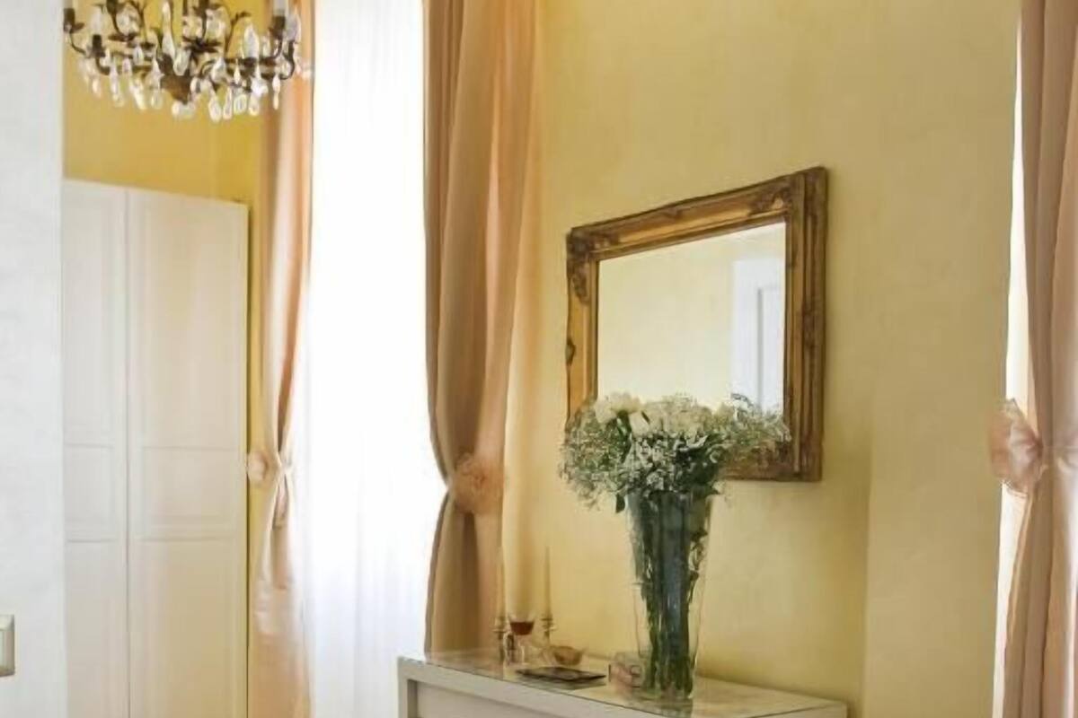 A bedroom with a bed, a vanity with a mirror, a chandelier, and a window with curtains.