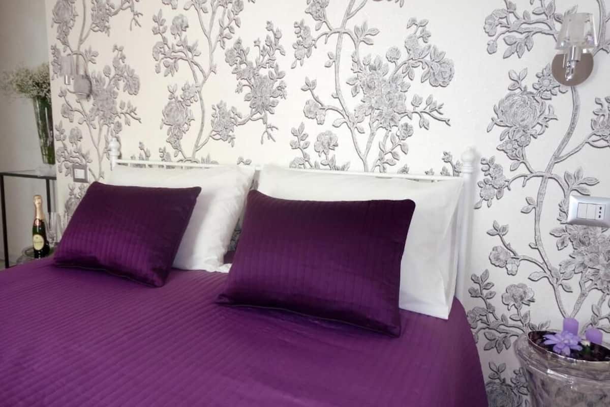 A bed with purple bedding, a white headboard, and a floral wallpaper pattern.