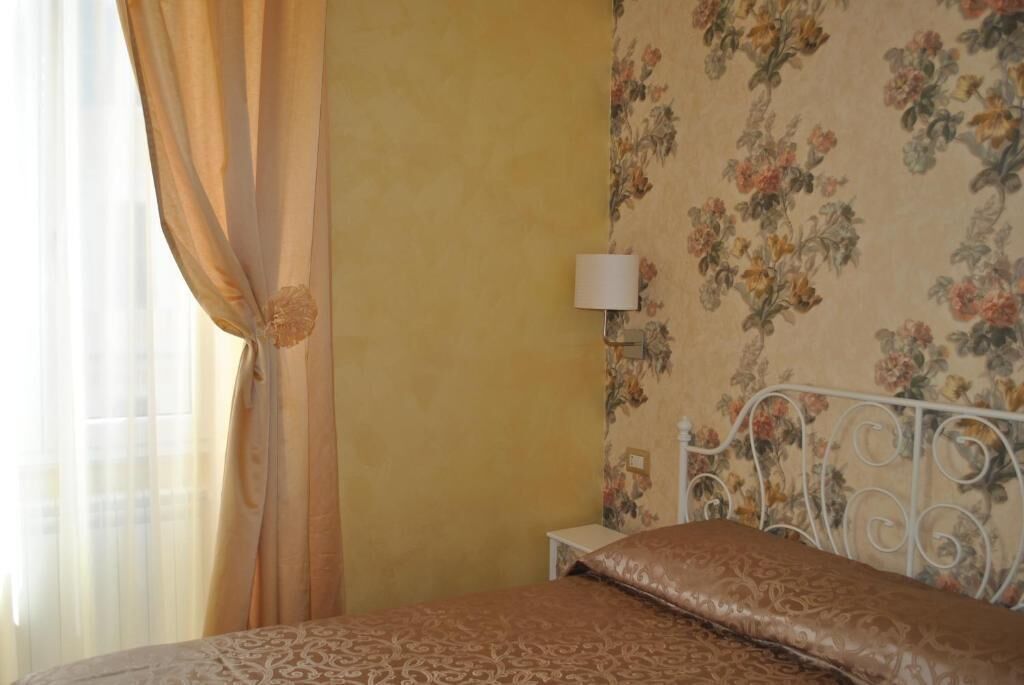 A bedroom with a bed, curtains, a floral wallpaper, and a nightstand with a lamp.