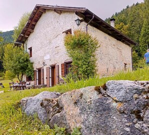 A rustic stone building with a sloped roof, wooden shutters, and a small porch with outdoor seating.
