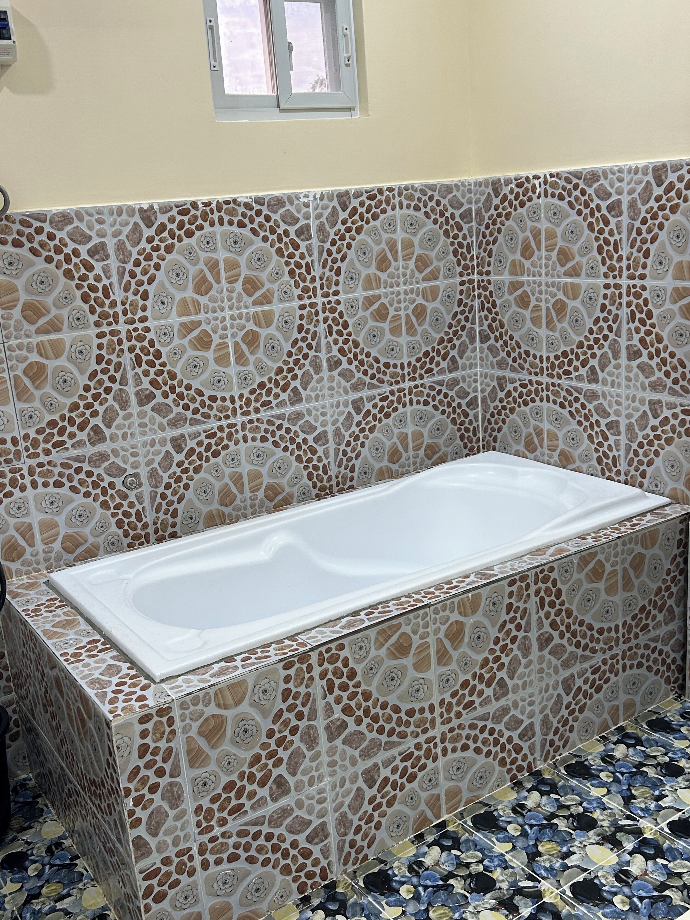 Un baño con una bañera rectangular, paredes con mosaicos y una ventana sobre la bañera.