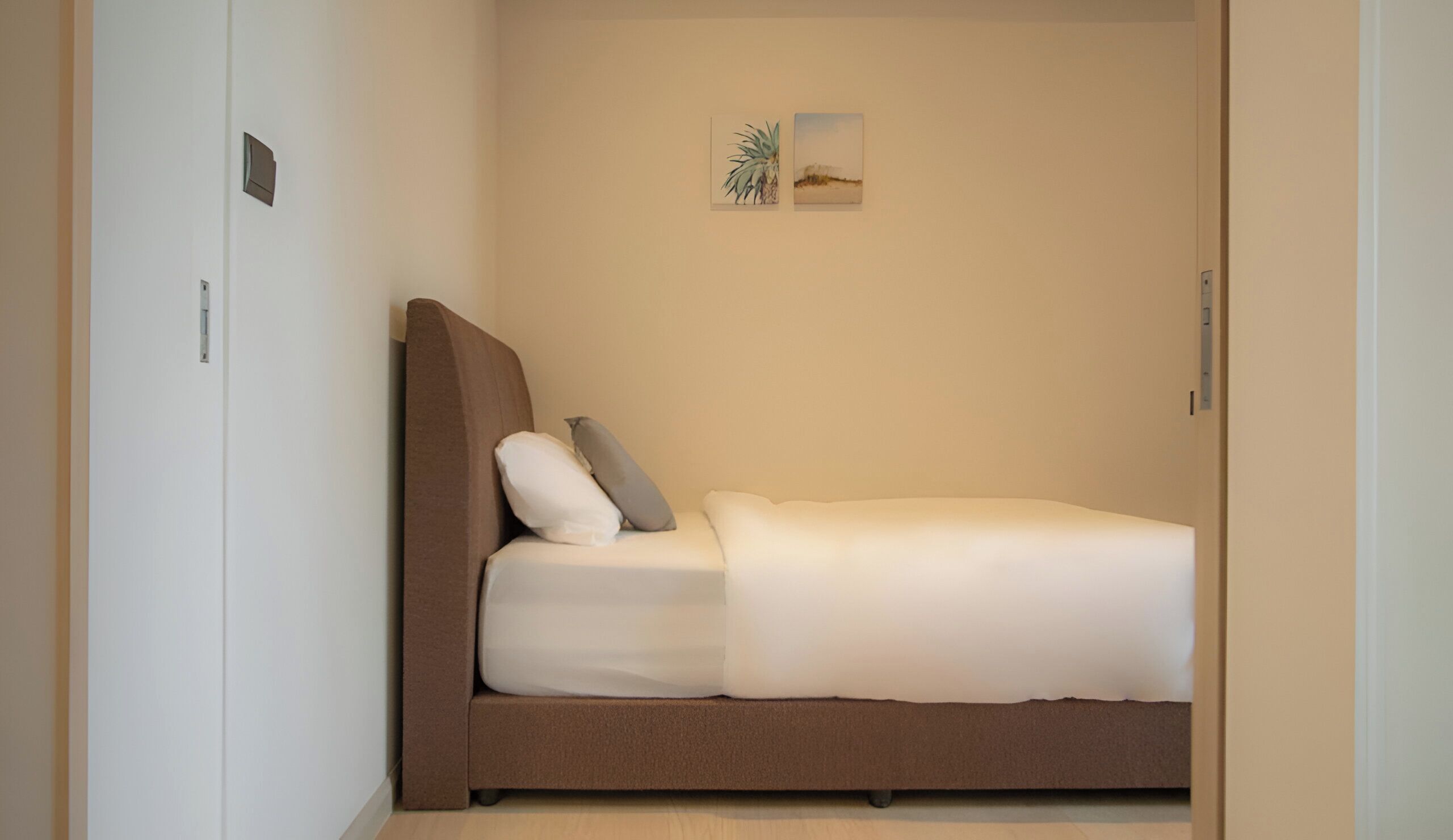 Une petite chambre avec un lit simple, une tête de lit marron et du linge de lit blanc. On y trouve deux interrupteurs fixés au mur et deux tableaux encadrés au-dessus du lit.