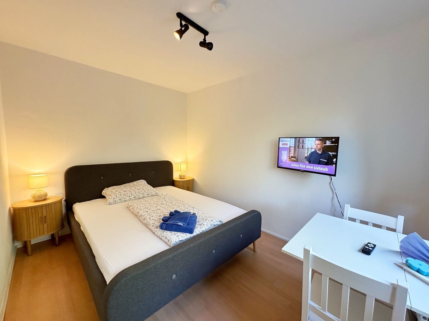 Ein Schlafzimmer mit einem Bett, Nachttischlampen, einem an der Wand montierten Fernseher und einem kleinen Tisch mit Stuhl.