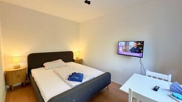 Ein Schlafzimmer mit einem Bett, Nachttischlampen, einem an der Wand montierten Fernseher und einem kleinen Tisch mit Stuhl.