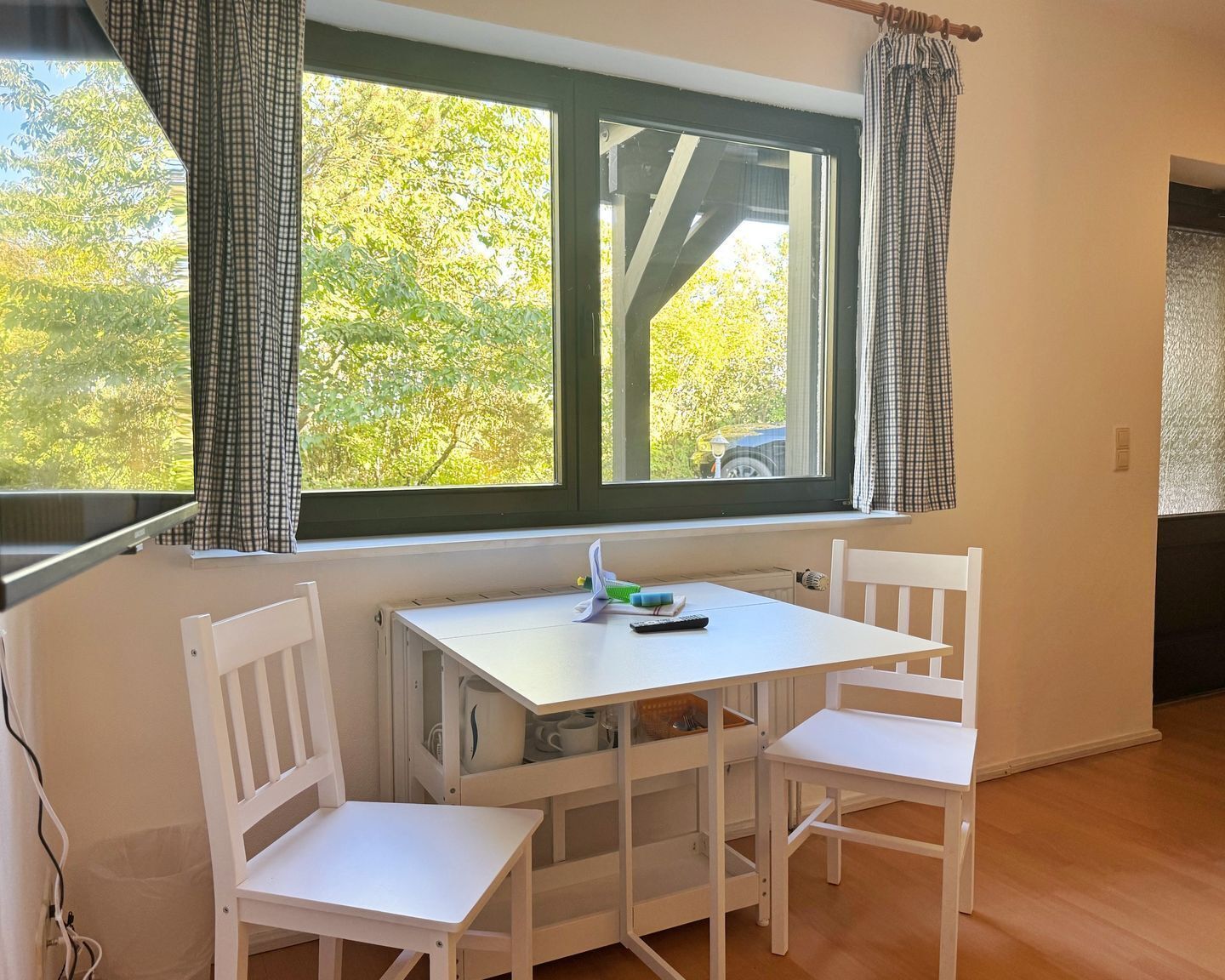 A small room with a white table and two white chairs, a window with curtains, and a view of trees outside.