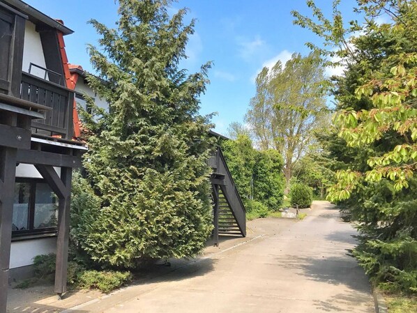 Un chemin mène à un bâtiment doté d’un escalier en bois et d’un grand arbre situé sur la gauche.