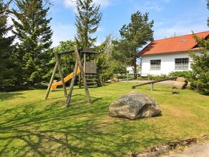 A playground with a wooden swing set and slide, a large rock, and a white building with a red roof in the background.