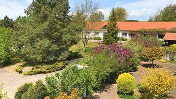 Ein angelegter Garten mit verschiedenen Bäumen und Sträuchern, eine gepflasterte Auffahrt und ein Gebäude mit rotem Ziegeldach.