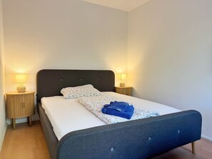 Un lit bien fait, avec une tête de lit sombre, deux tables de chevet avec des lampes et un plancher en bois.