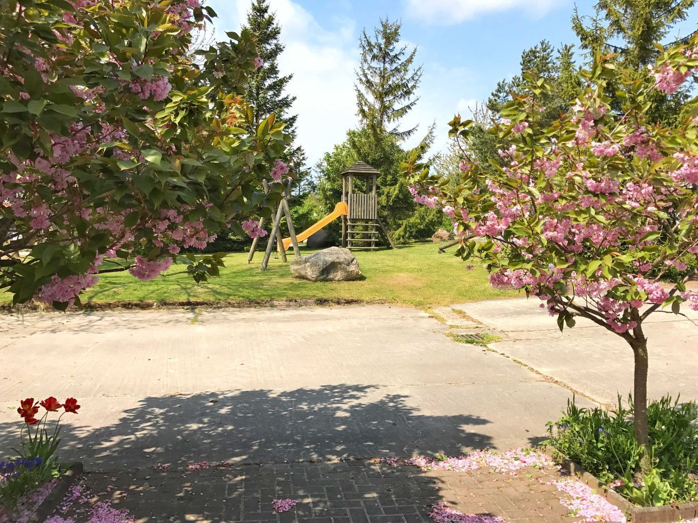 Ein Spielplatz mit Rutsche und Klettergerüst, umgeben von blühenden Bäumen und einem gepflasterten Weg.