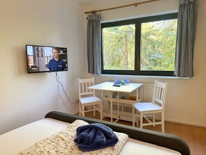 A hotel room with a bed, a TV mounted on the wall, a window with curtains, and a small table with chairs.