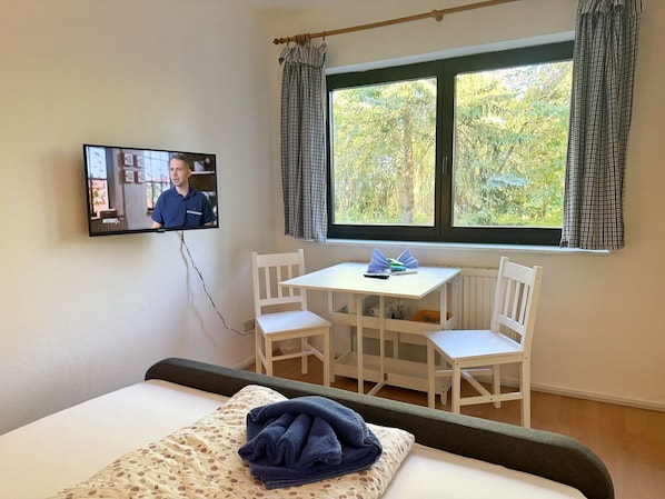 Hotellihuone, jossa on sänky, seinälle asennettu televisio, ikkuna verhoilla sekä pieni pöytä ja tuolit.