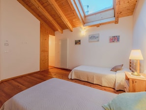 Quarto com duas camas, vigas de madeira no teto, claraboia, radiador e mesa de cabeceira com abajur.