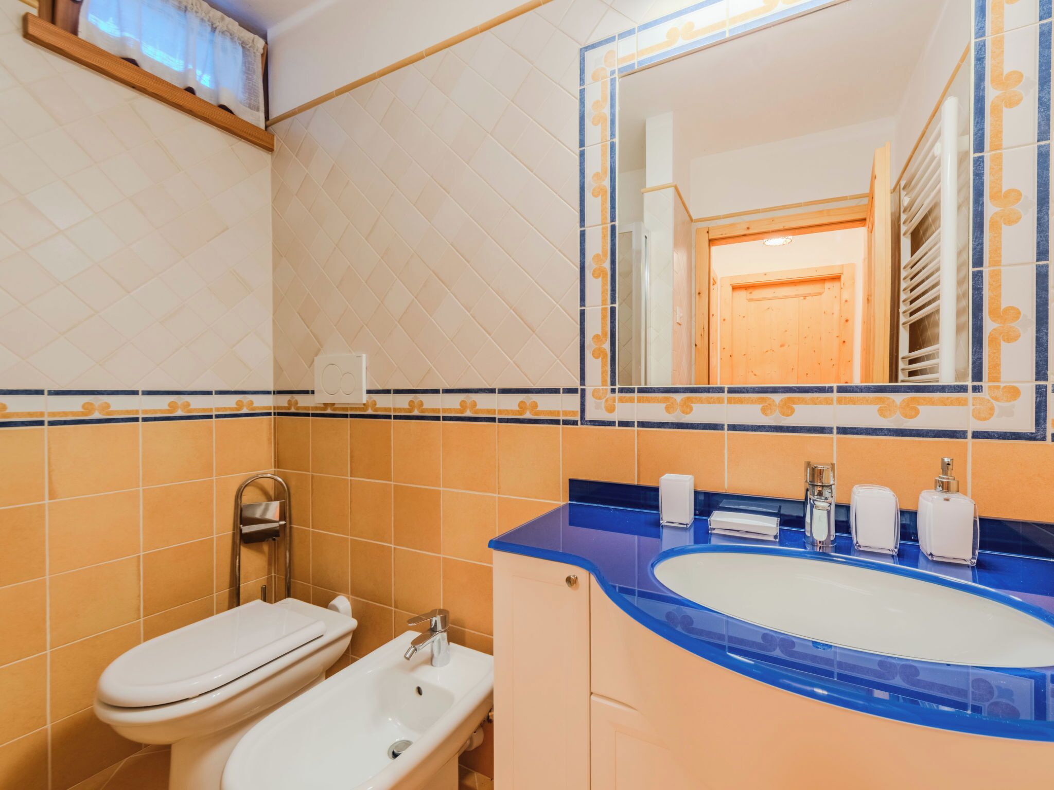 Ein Badezimmer mit Toilette in Weiß, einem Waschbecken mit blauer Ablage und einem Spiegel, der eine Holztür reflektiert.