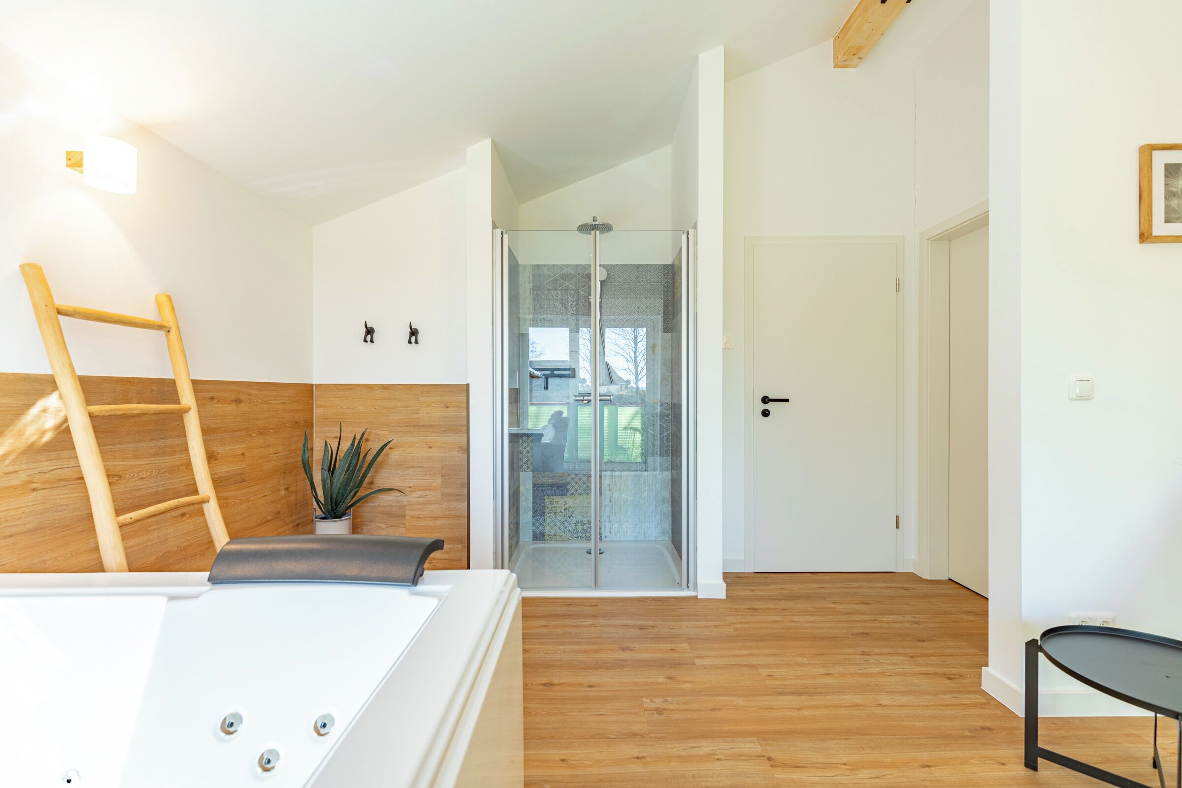 A modern bathroom with a wooden floor, a white bathtub, a shower with glass doors, and a small potted plant.