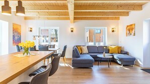 A modern living room with a wooden ceiling, a grey sectional sofa, a dining table with chairs, and a small round table with a vase of flowers.