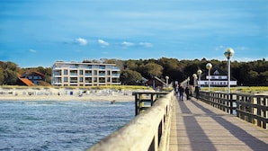 A wooden boardwalk leading to a beach with a modern building and a house in the background. - Vacation apartments near the beach on the pier (Graal-Müritz)