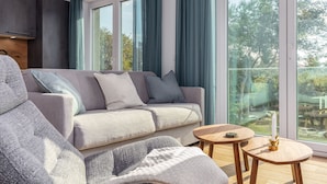 A modern living room with a grey sofa, a wooden coffee table, and a view of the outdoors through large windows. - Vacation apartments near the beach on the pier (Graal-Müritz)