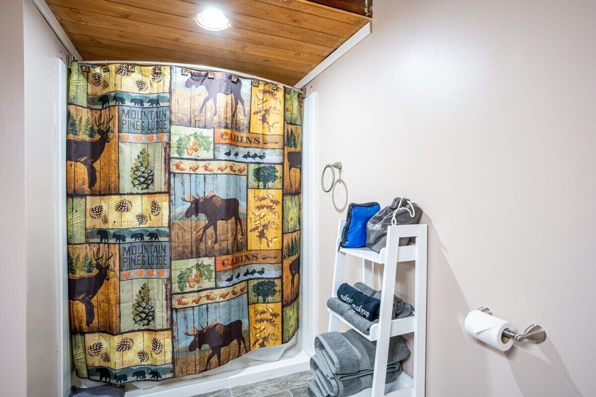 A bathroom with a wooden ceiling, a shower curtain with wildlife and cabin-themed designs, a towel rack with towels, and a toilet paper holder.