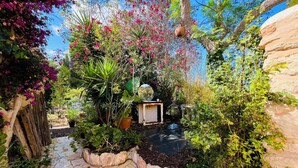 Un sendero de piedra conduce a una estructura de madera rústica, rodeada de abundante follaje y plantas con flores.