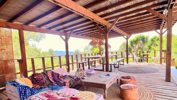 Un área de estar al aire libre techada con un piso de madera, bancos y una mesa con un tapete tejido.