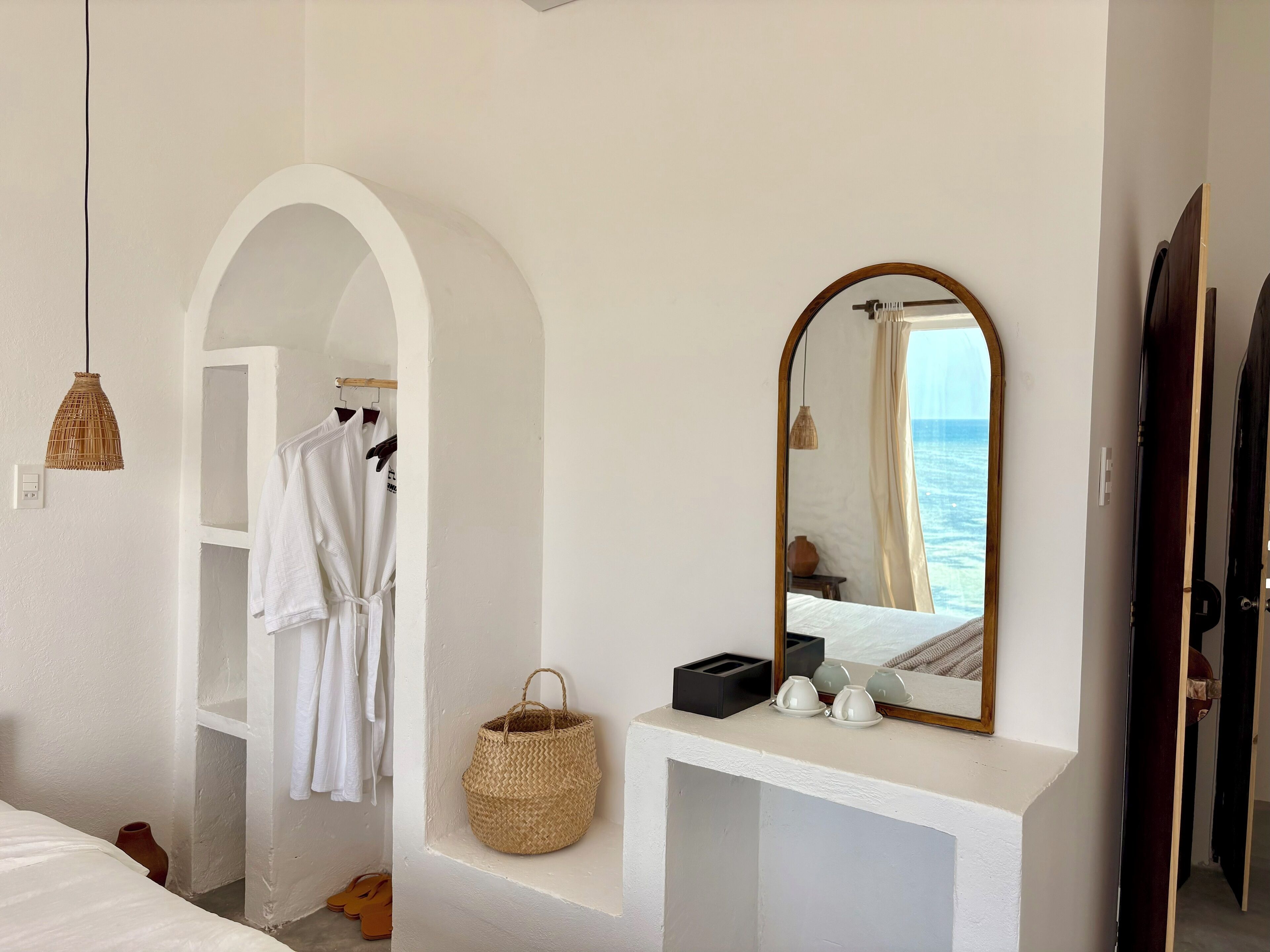 A bedroom with a large arched doorway, a bedside table with a mirror, and a view of the sea through the window.
