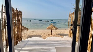 En vy över stranden genom en glasdörr, med en sandstig som leder ner till vattnet, ett parasoll med halmtak och ett trästaket.