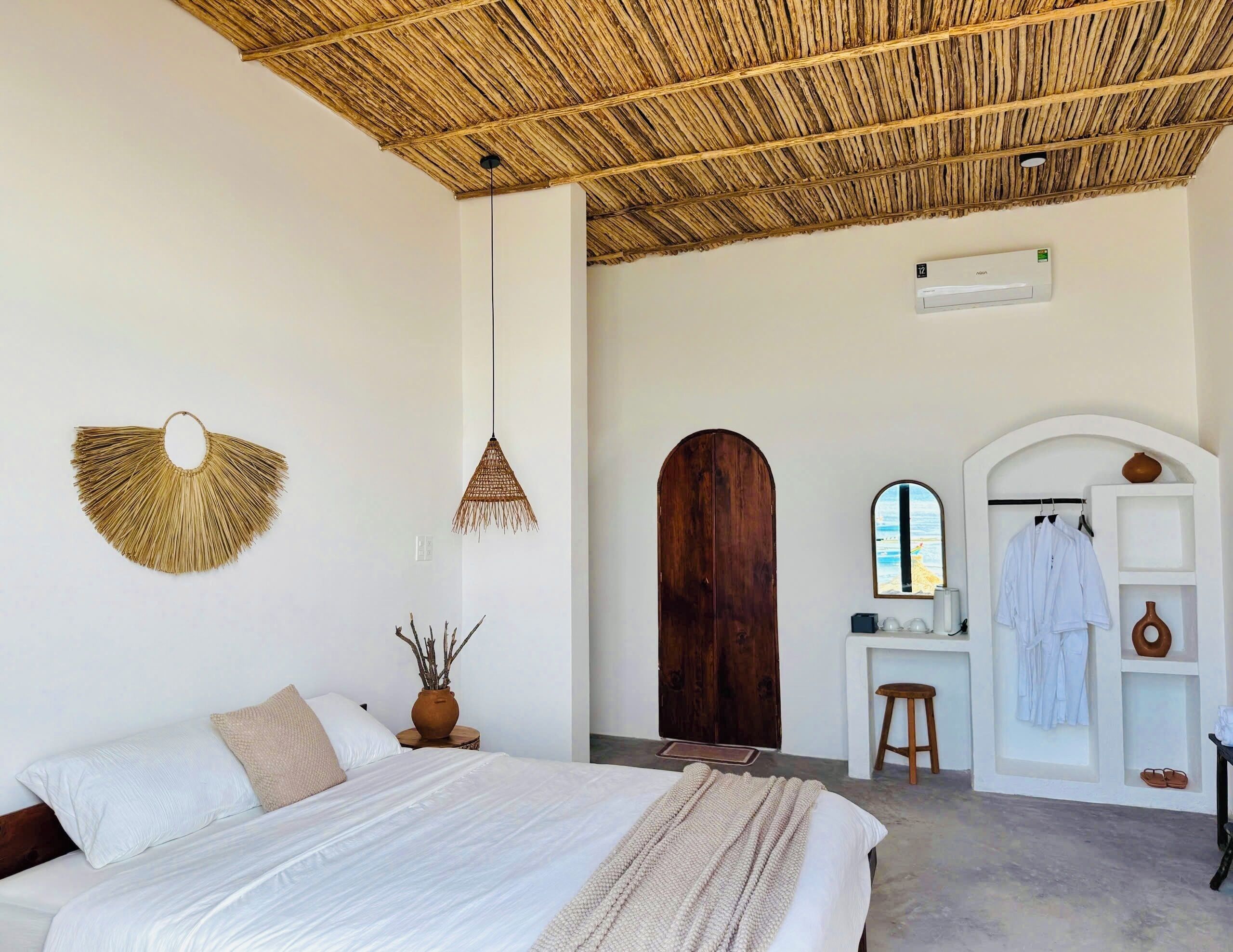 A bedroom with a bed, a hanging light, a wooden door, a mirror, and a shelf with clothes and a vase.
