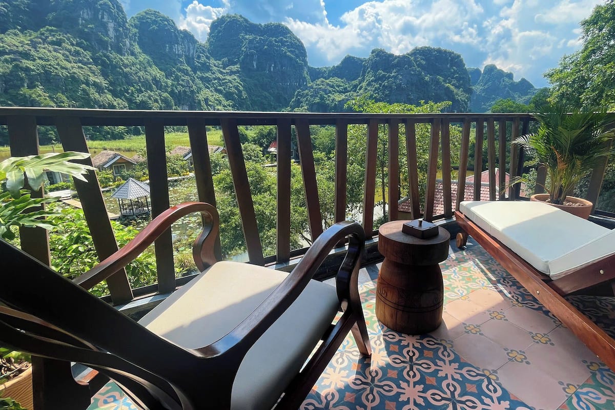 A balcony with two lounge chairs, a wooden table, and a view of lush greenery and mountains.