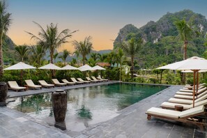 Un espace aménagé au bord de la piscine, avec des chaises longues et des parasols, entouré de palmiers et dominé par un paysage montagneux.