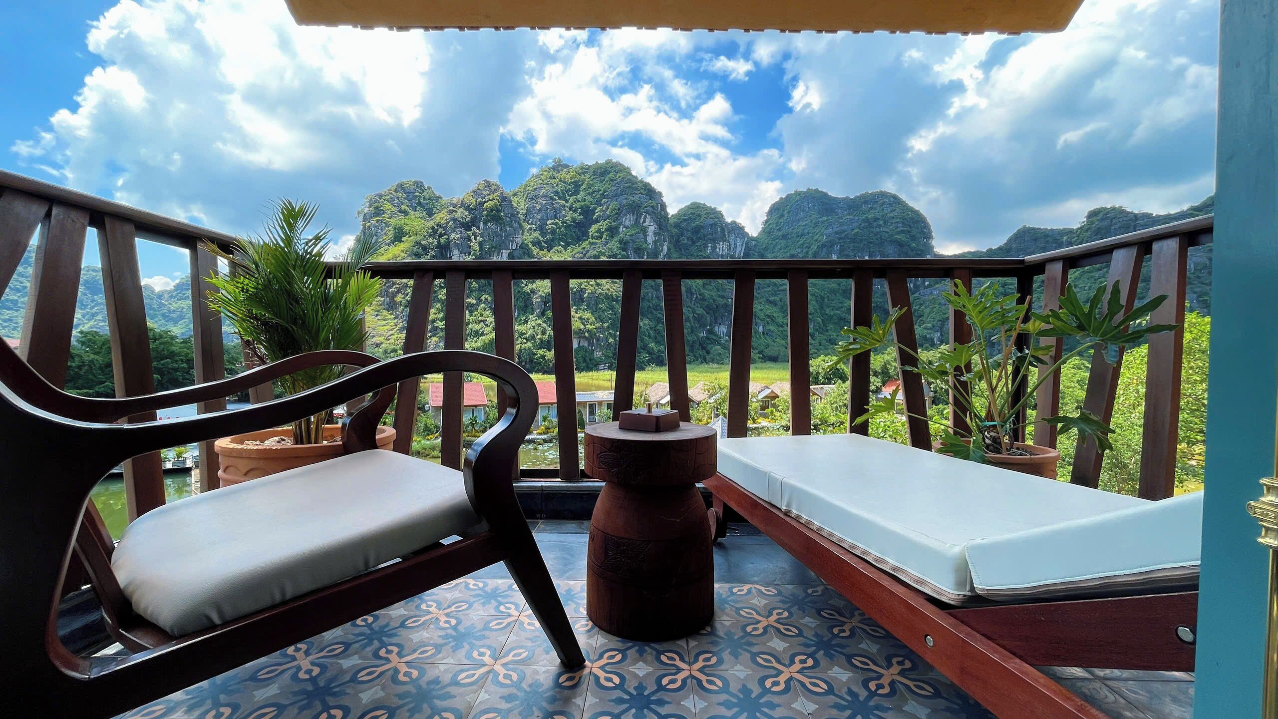 Un balcón con vistas a un paisaje montañoso, una silla de madera, una mesita redonda y un colchón.