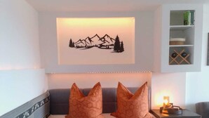A hotel room with a bed, two orange pillows, a wall-mounted artwork of mountains, a bedside table with a candle, and a shelf with books and a basket.