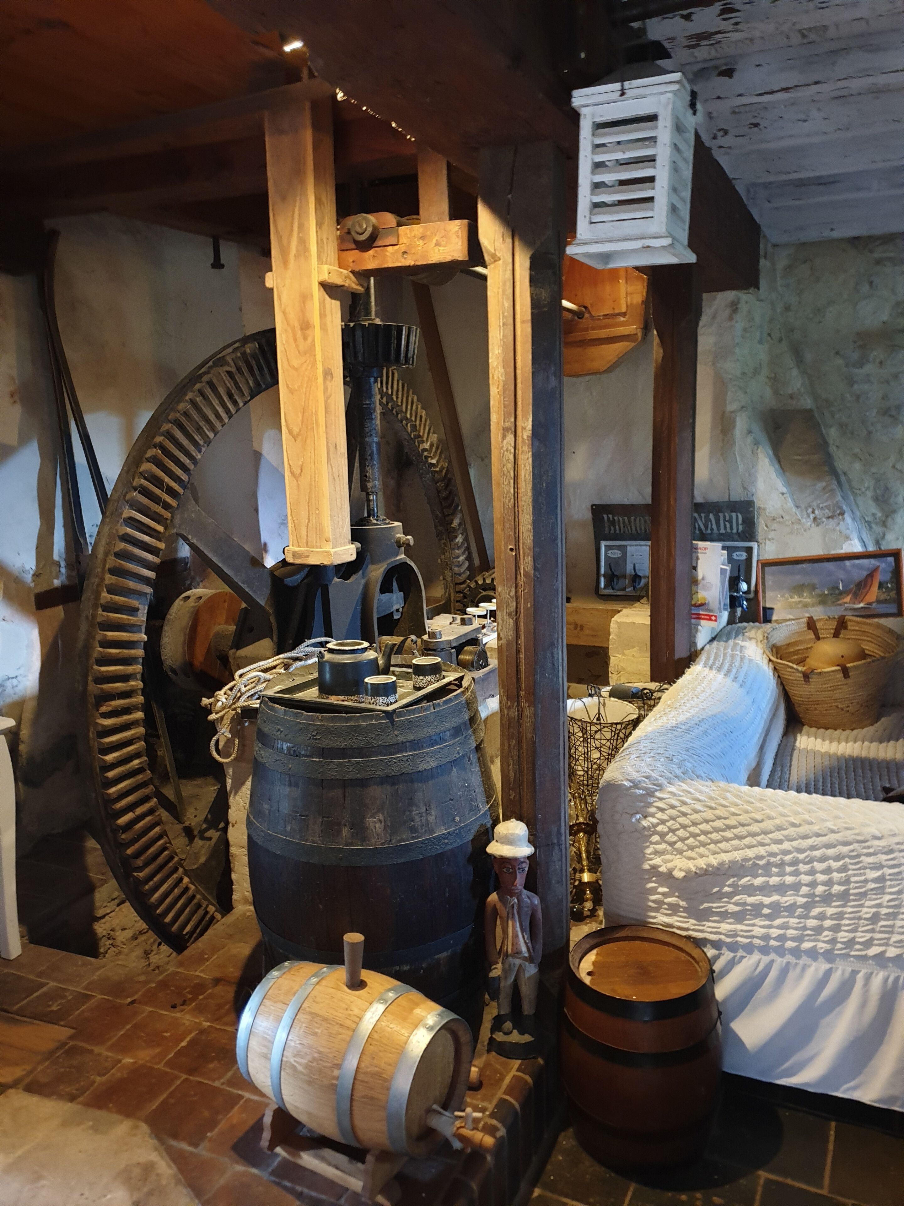 Une pièce rustique avec une grande roue à eau en bois, un lit recouvert d’une courtepointe blanche et un mur en pierre.