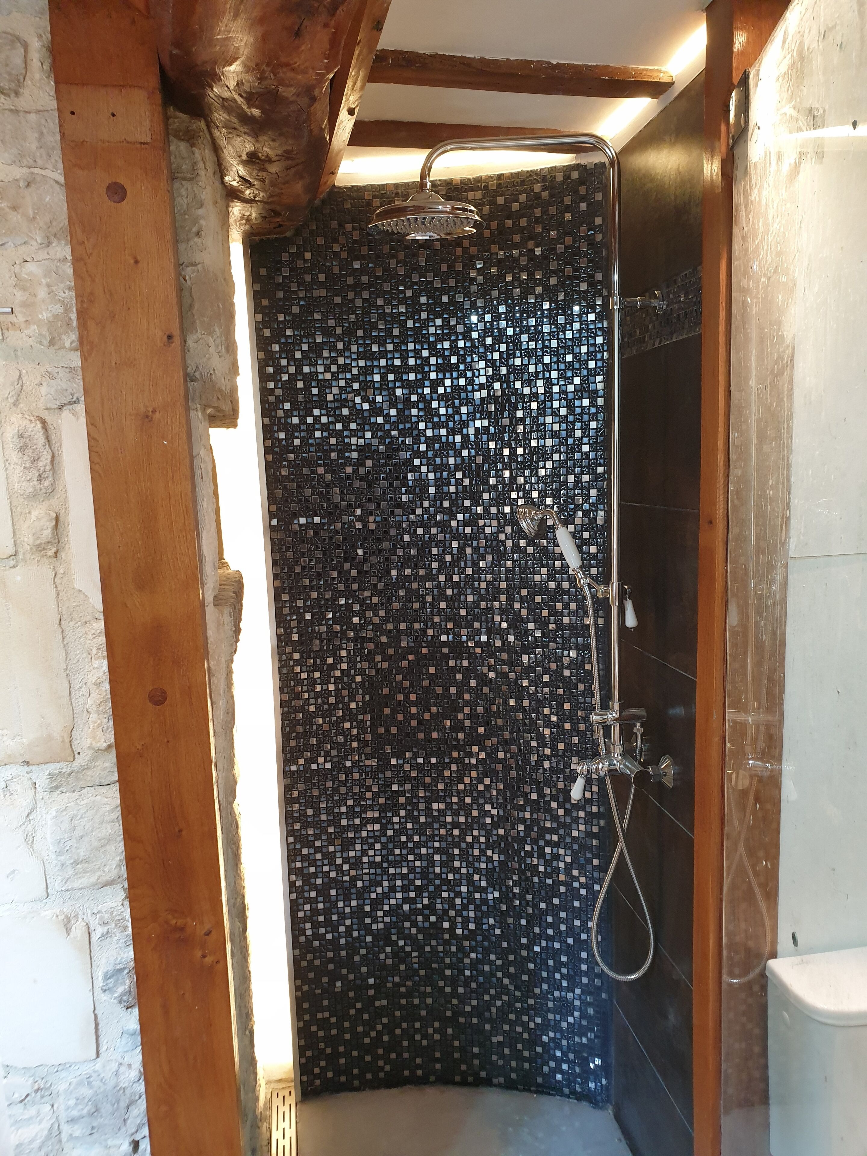 Una ducha con pared de mosaicos, vigas de madera y un inodoro blanco.
