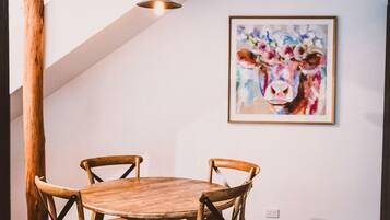 A dining area with a round wooden table and four wooden chairs. A framed painting of a cow is on the wall.
