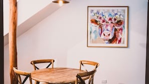 A dining area with a round wooden table and four wooden chairs. A framed painting of a cow is on the wall.