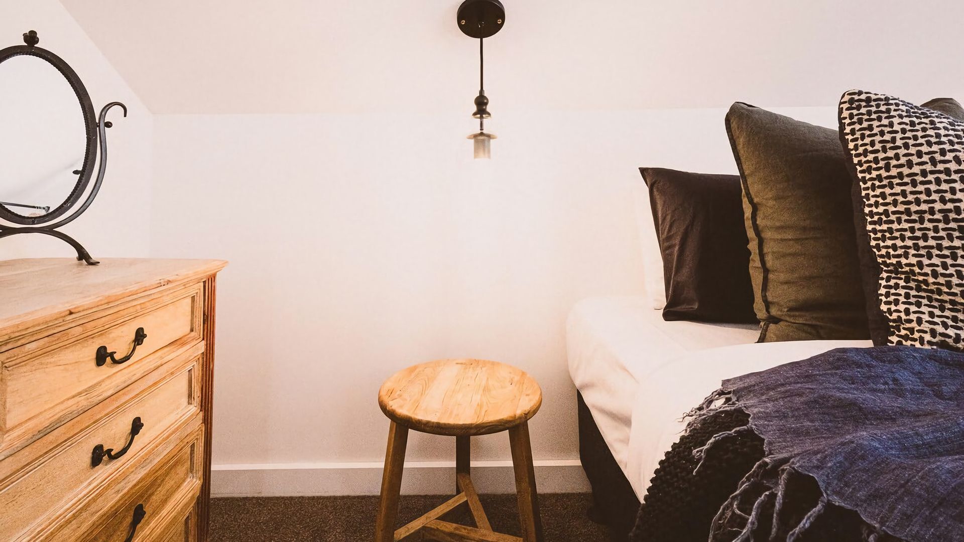 A bedroom with a wooden dresser, a stool, a bed with pillows, and a hanging light fixture.