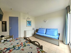 A room with a bed, a sofa, a dining table, chairs, a ceiling fan, a wall-mounted light, a door, and a window with curtains.